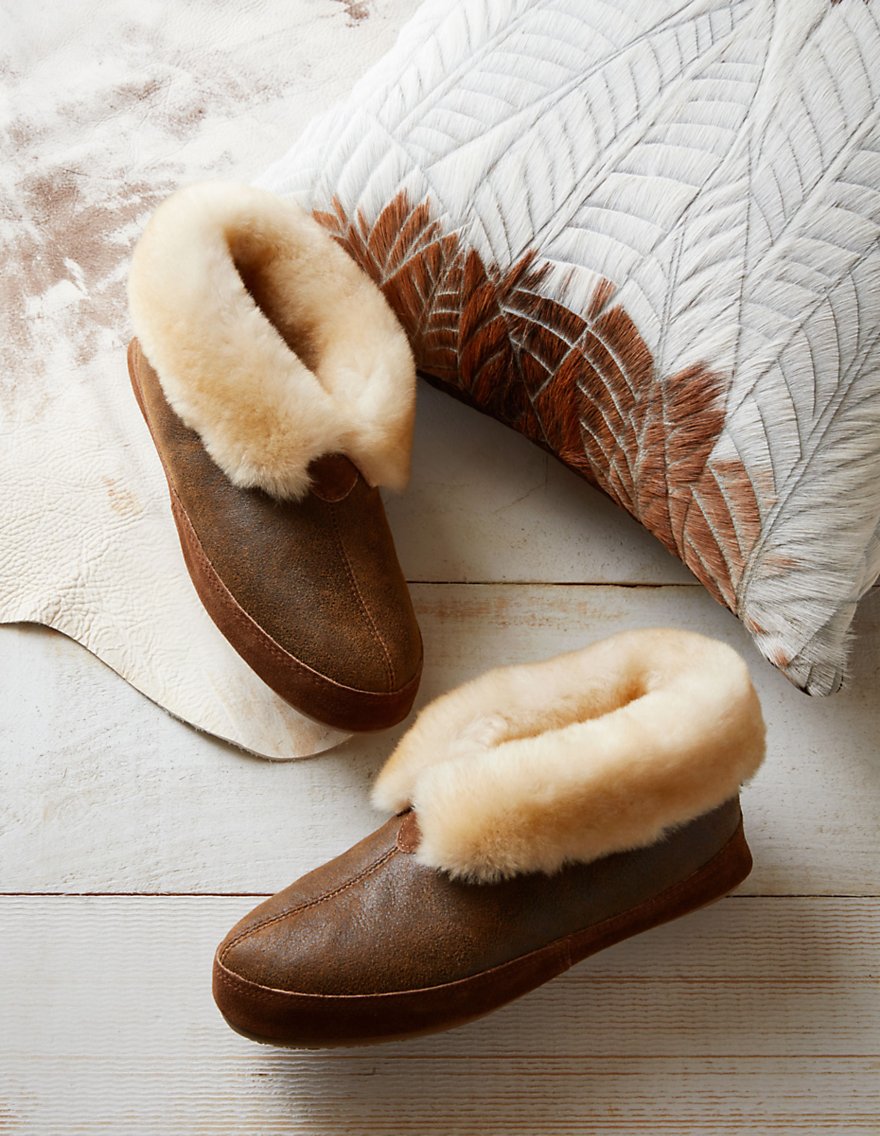 Men's Ethan sheepskin slippers on a wood and leather background with a cowhide pillow